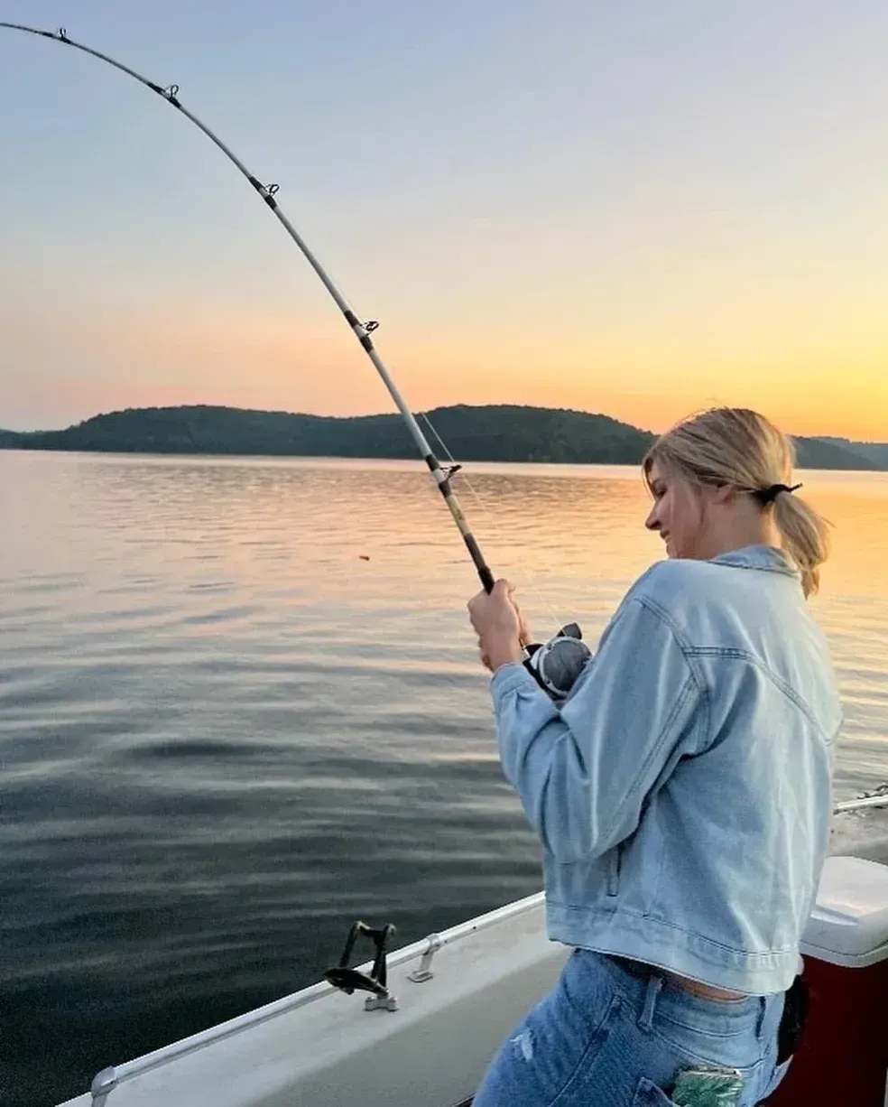 Woman fishing on a boat at sunset, holding a fishing rod, calm water, light blue and orange sky.