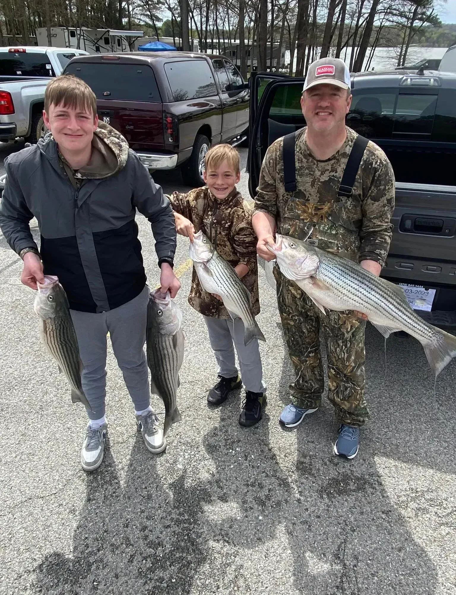 Three people holding fish near vehicles. The person on the right is smiling.