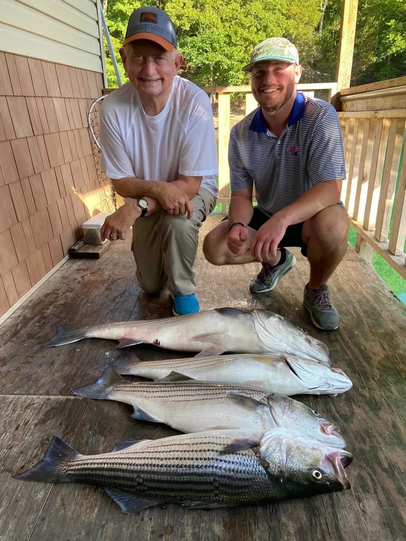 Two men pose with four striped bass on a wooden deck. The older man kneels, and the younger man squats.