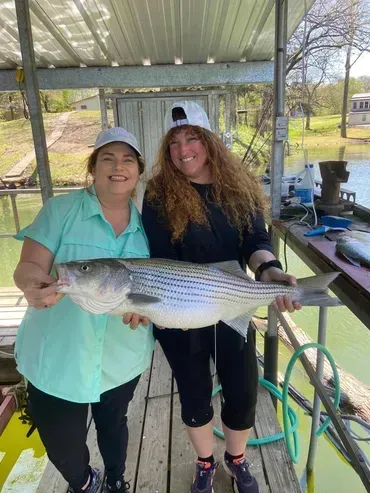 Two women holding a large striped fish on a dock, smiling.