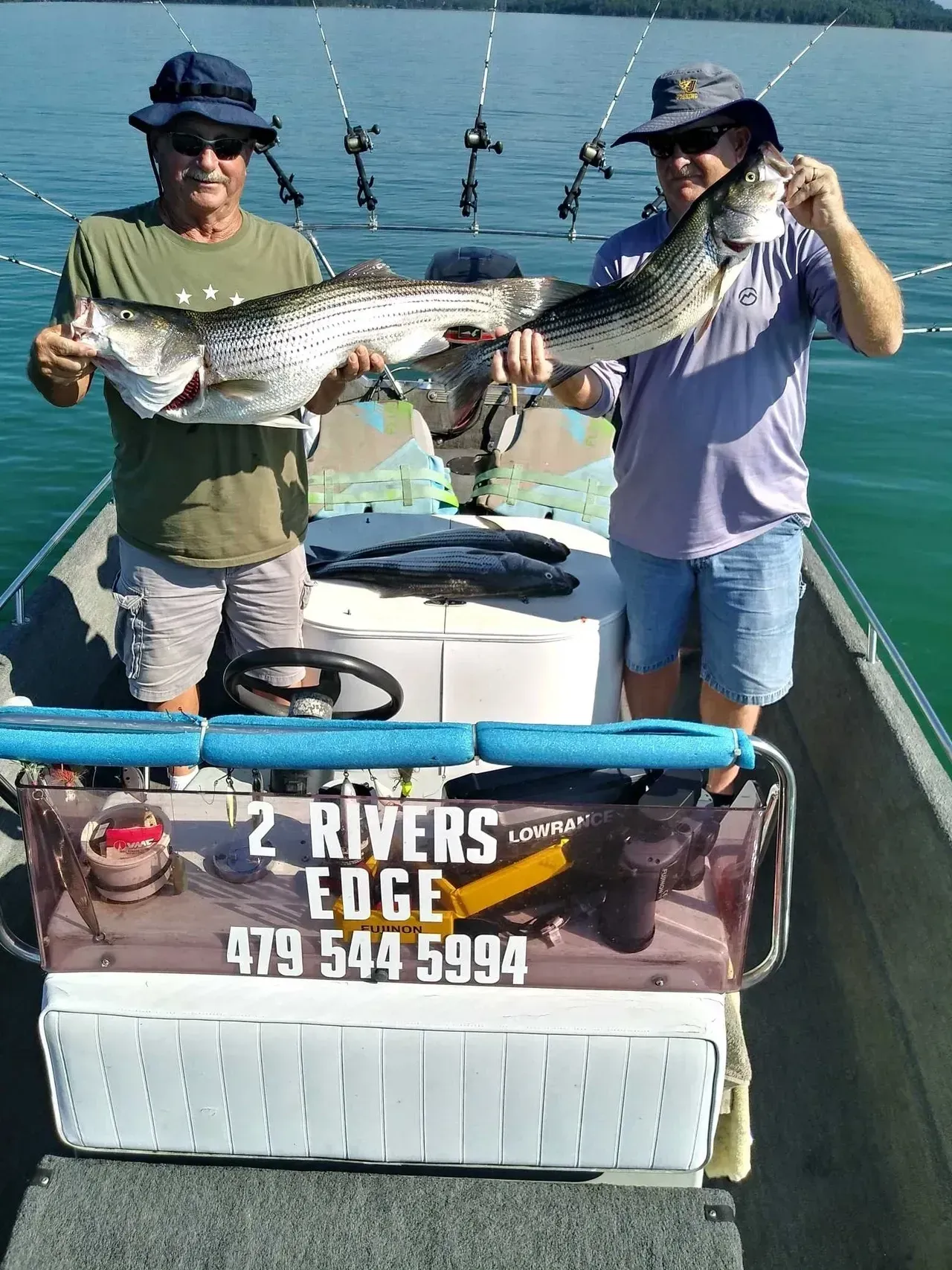 Two men on a boat holding large fish. Lake setting, blue water, sunny day.