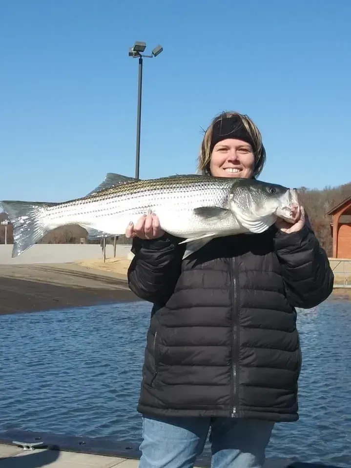 Woman in black jacket holds a striped bass fish at a waterside location on a sunny day.