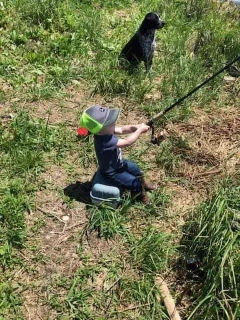 A child fishing, sitting on a tackle box. A dog sits in the grass nearby.