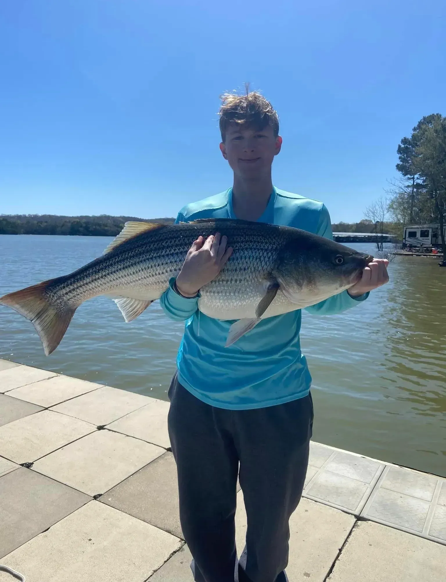 Teen holding a large striped fish, standing on a dock by water.