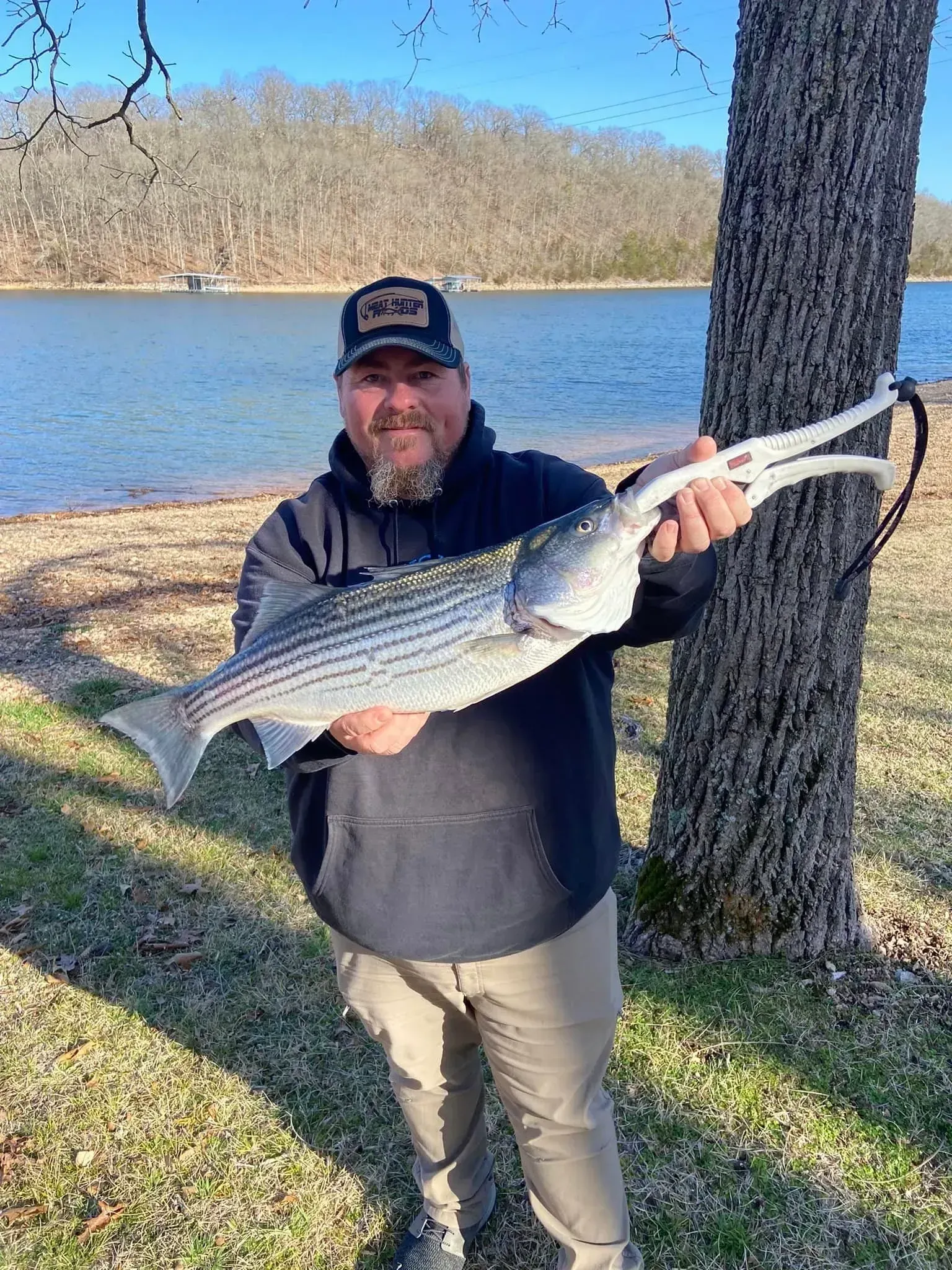 Man holding large striped fish near a lake; he's wearing a hat, hoodie, and khakis.