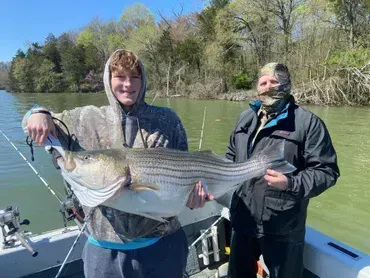Two people on a boat holding a large striped bass fish. The boy smiles, the other man's face is covered.