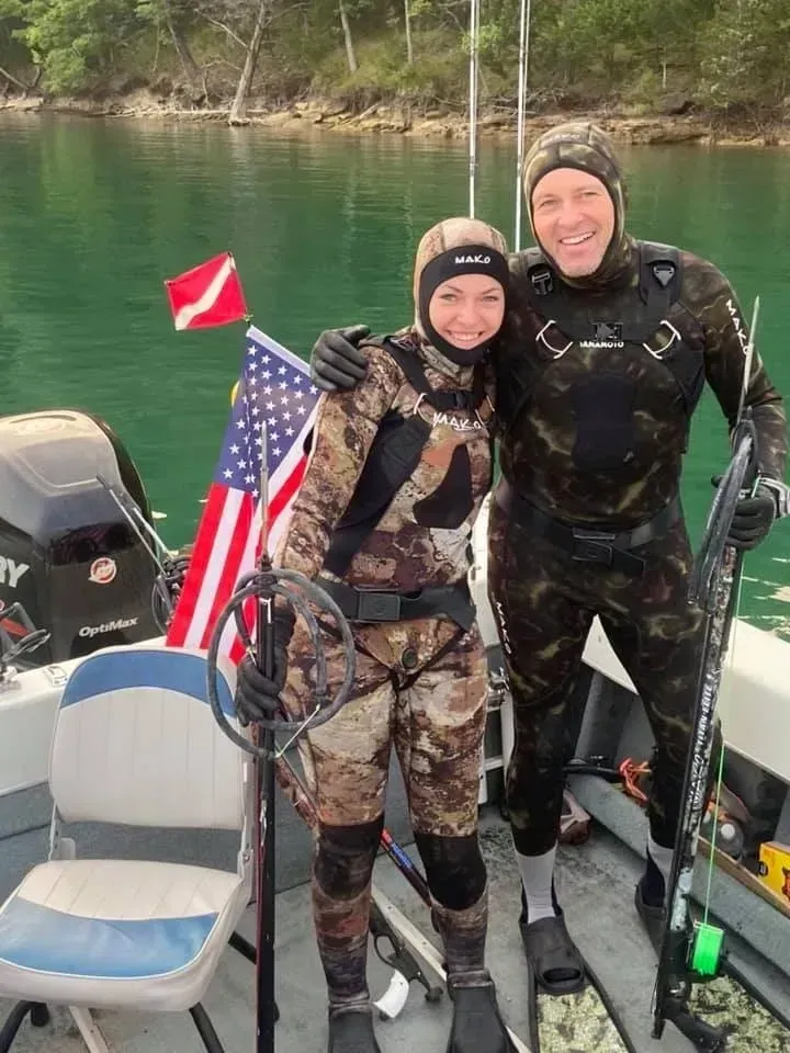 Two divers in camouflage wetsuits on a boat, holding dive gear, posing next to an American flag and serene water.