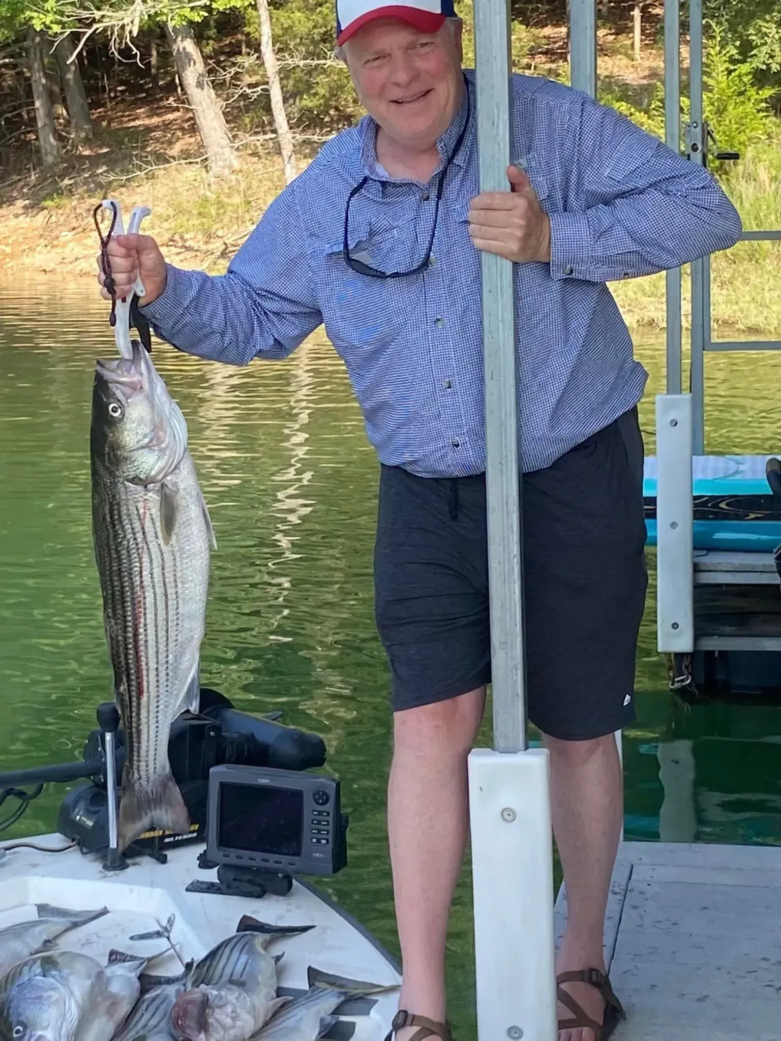 Man on dock holding up a large striped fish; other fish laid out. Water and trees in the background.