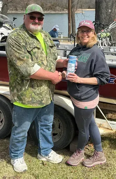 Man in camo shakes hands with woman holding a product; they stand by a red boat.