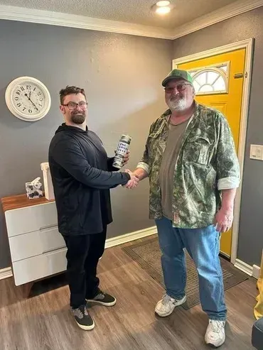 Two men shaking hands in a room. One holds a spray can. The room has a yellow door and a wall clock.