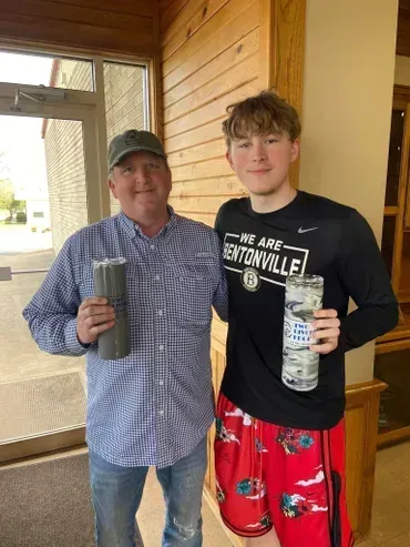 Man and young person smiling, holding tumblers, standing near a wooden wall, and door.