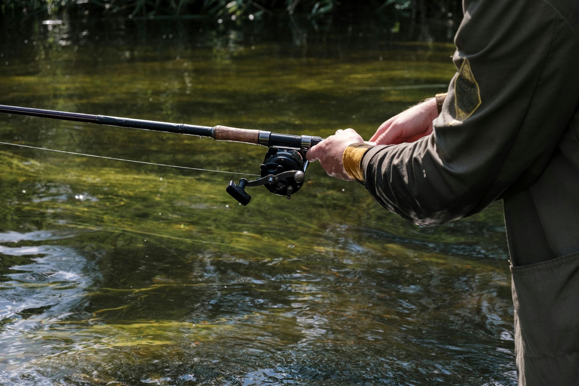 Person fishing in a stream, holding a fishing rod over flowing water with green vegetation.