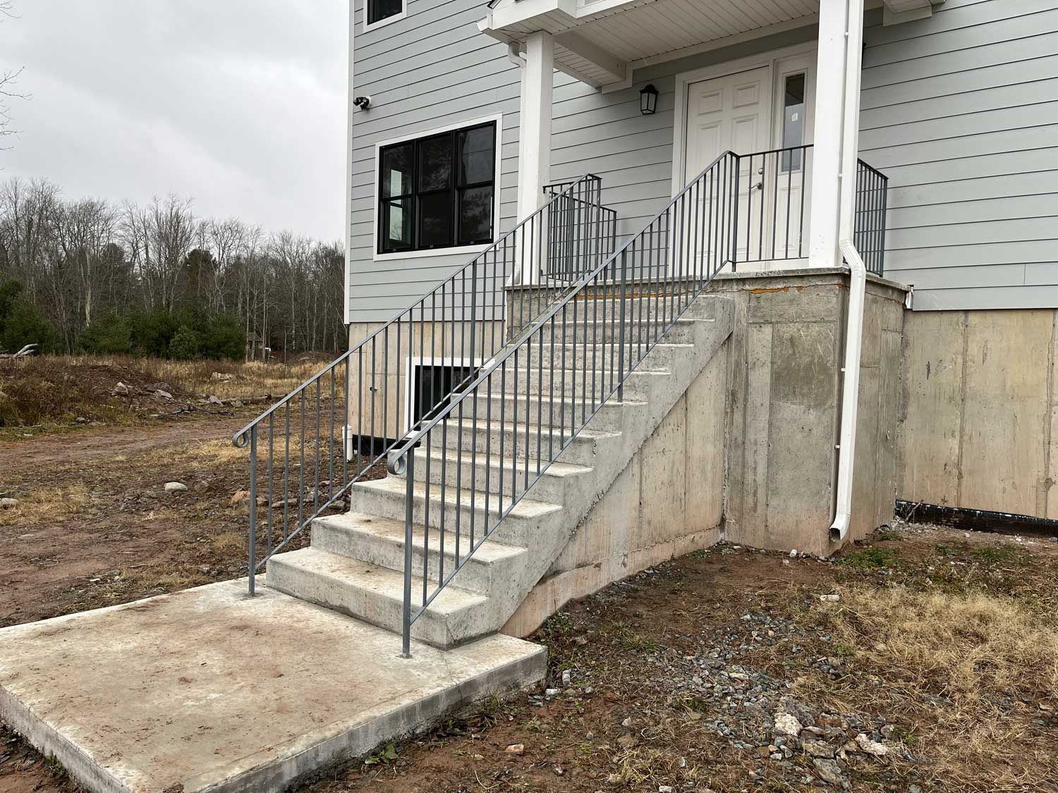 Concrete stairs with metal railings leading up to a house entrance. Cloudy sky, dirt ground.