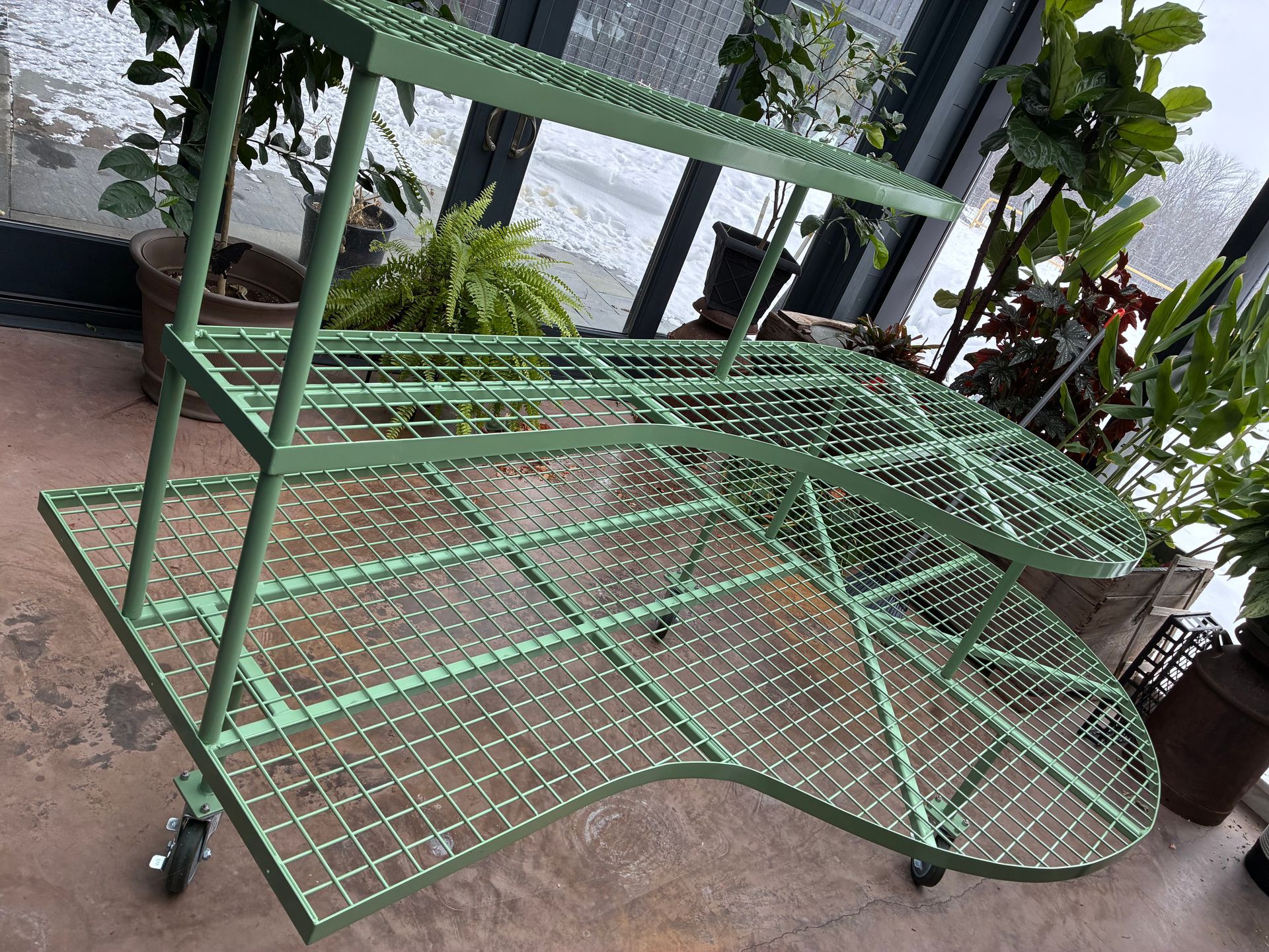 Green metal plant stand with three tiers, on wheels, indoors.