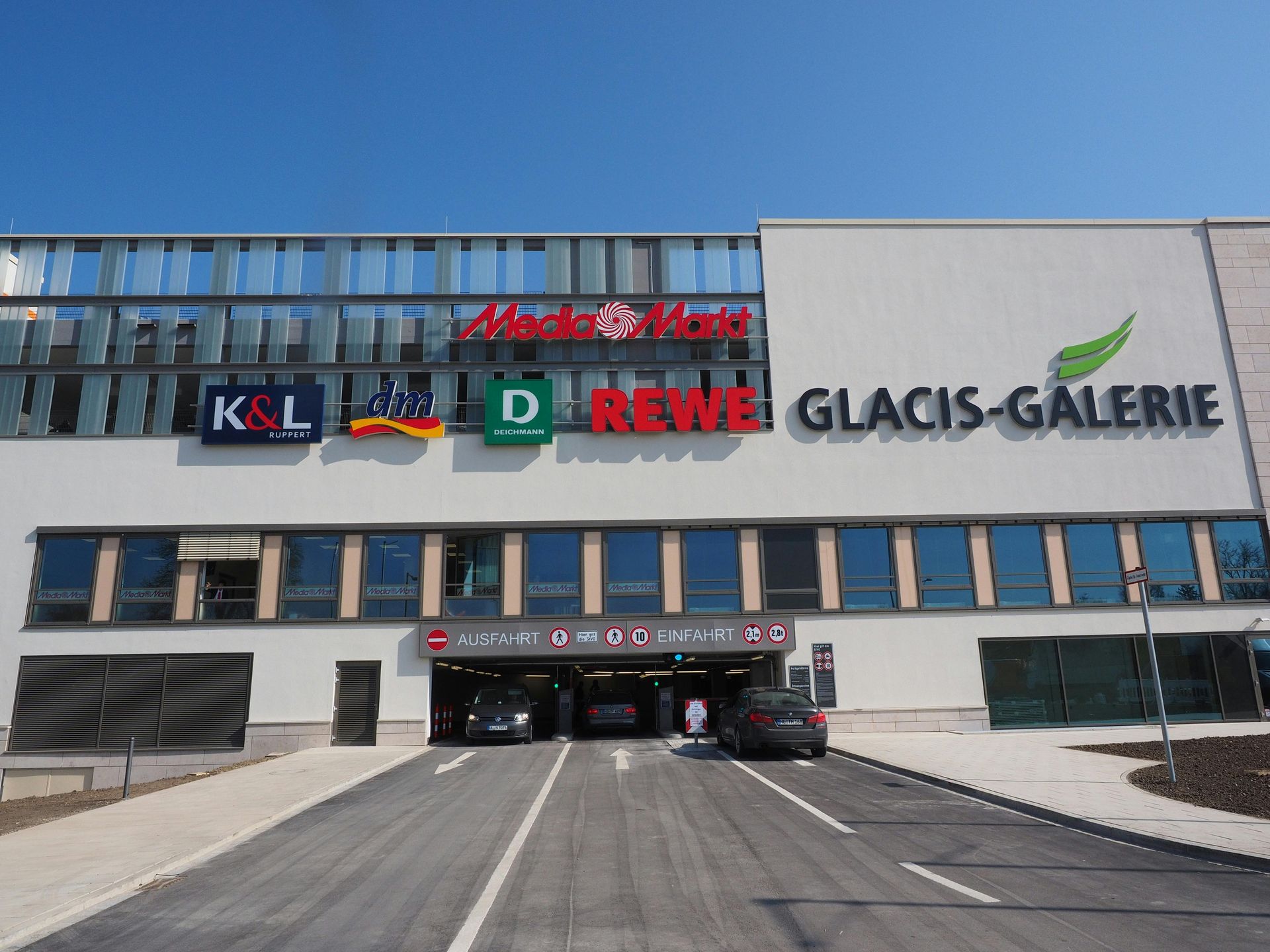 Exterior of Glacis-Galerie shopping center with multiple store signs: Media Markt, K&L, REWE. Parking entrance visible below.