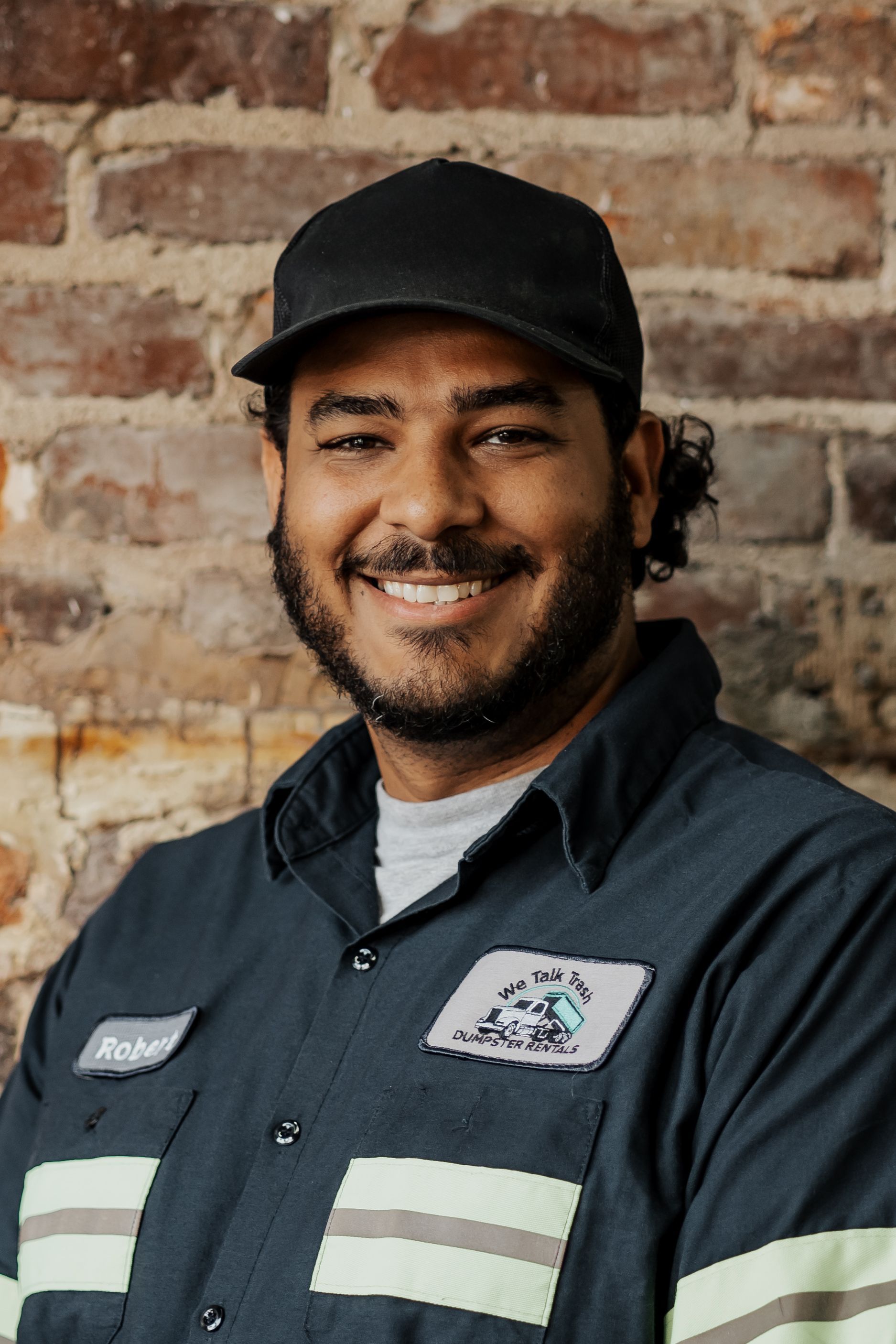 A man wearing a hat and a shirt is smiling in front of a brick wall.