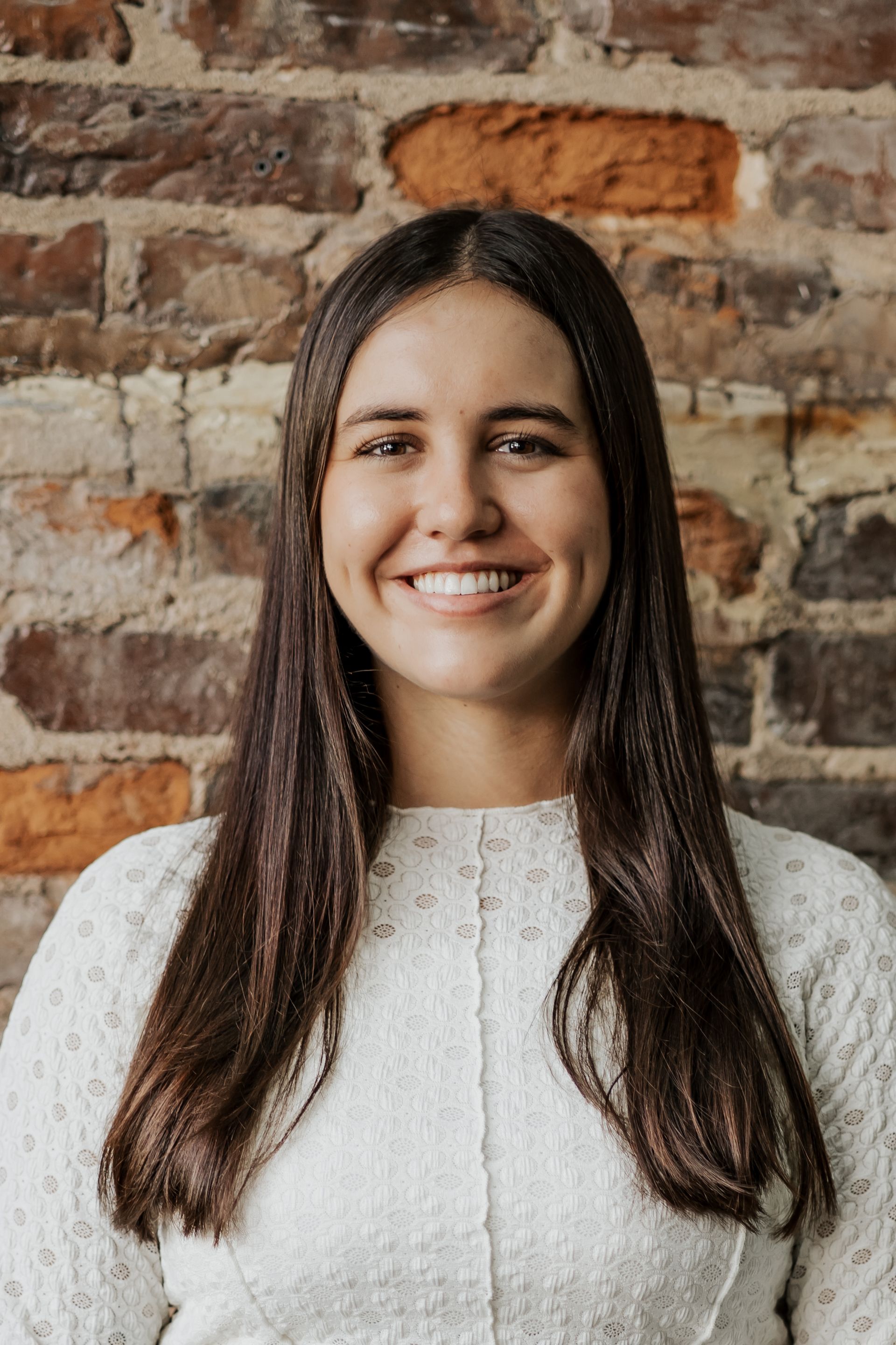 A woman in a white sweater is smiling in front of a brick wall.