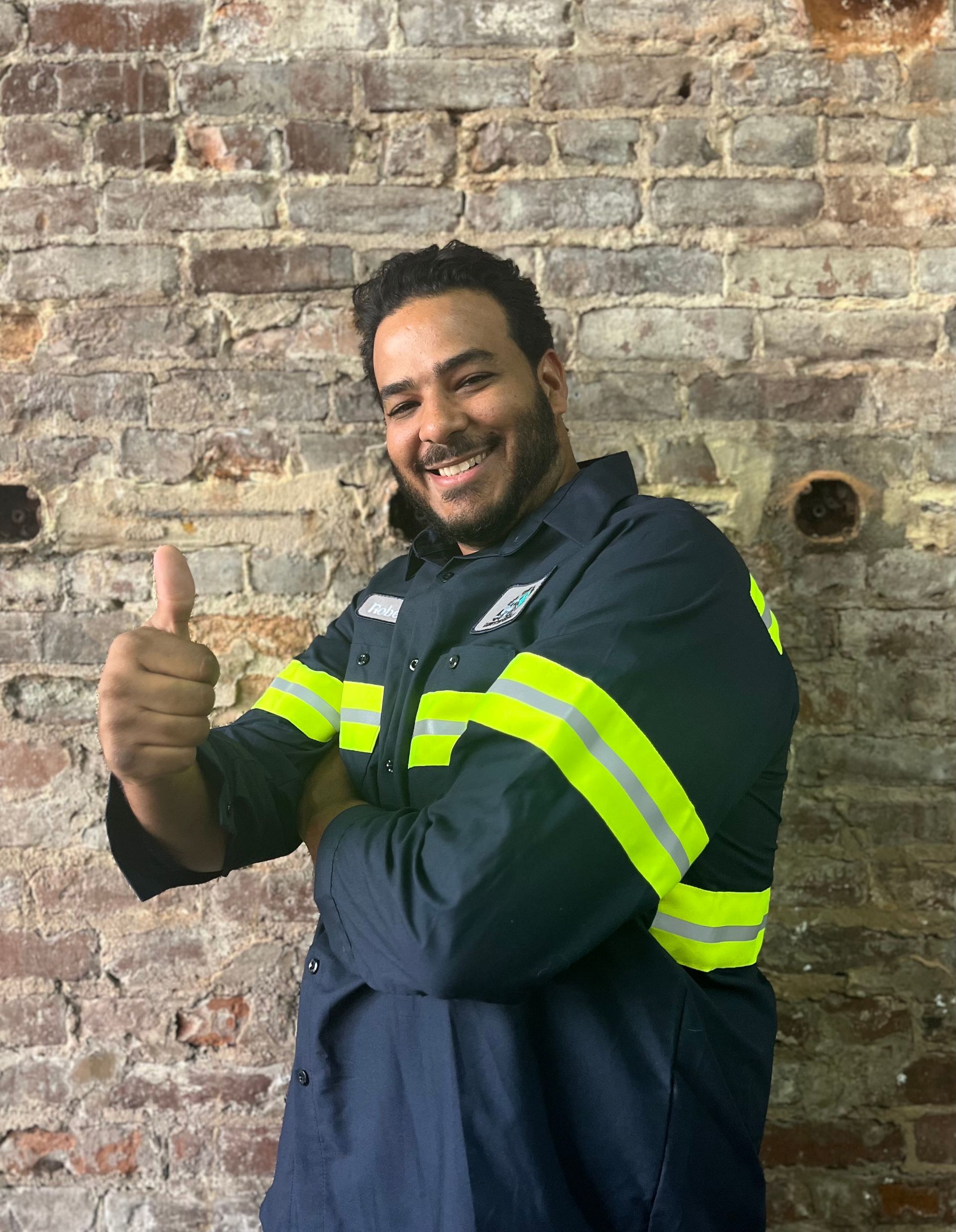 A man in a blue jacket is giving a thumbs up in front of a brick wall.