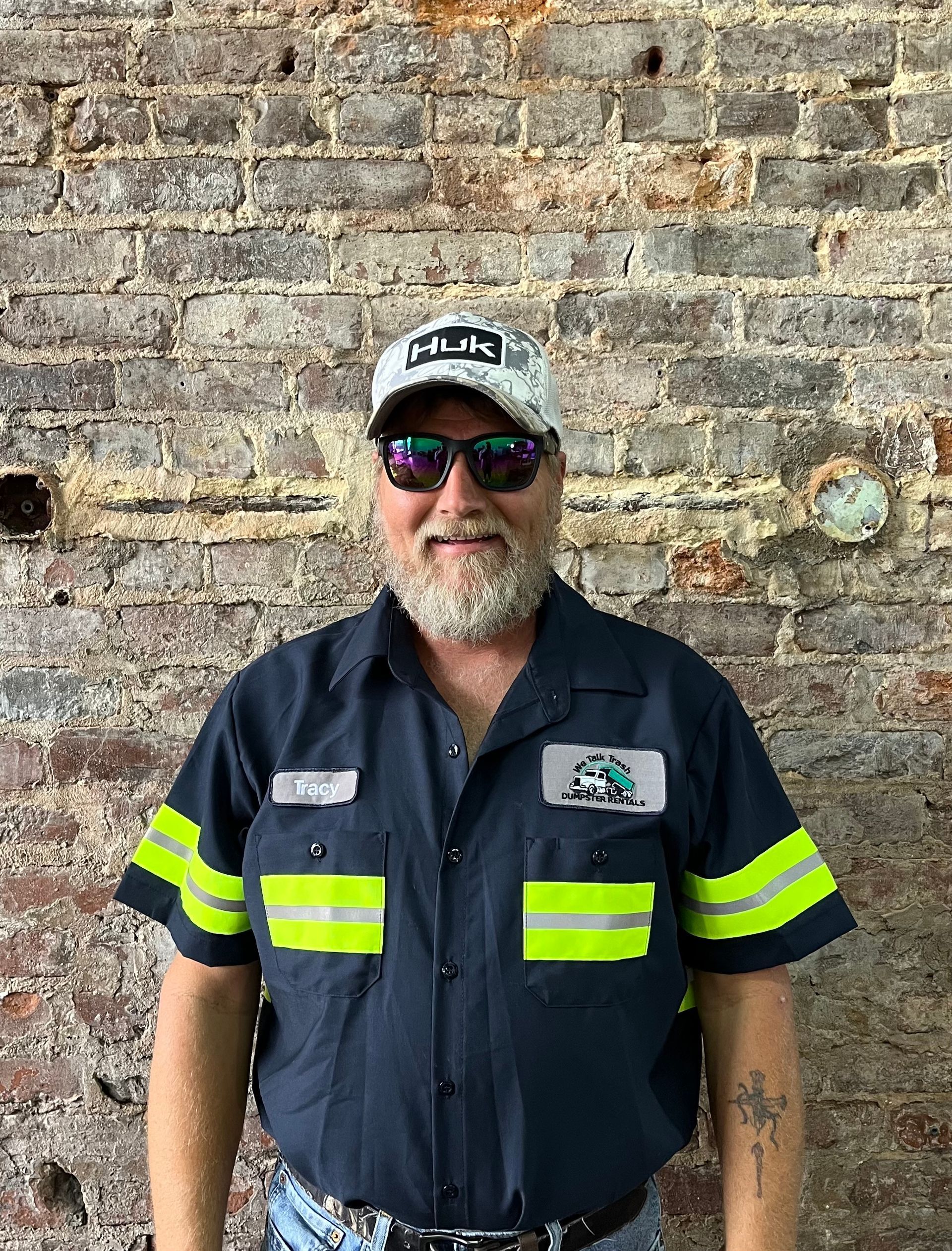 A man wearing sunglasses and a hat is standing in front of a brick wall.