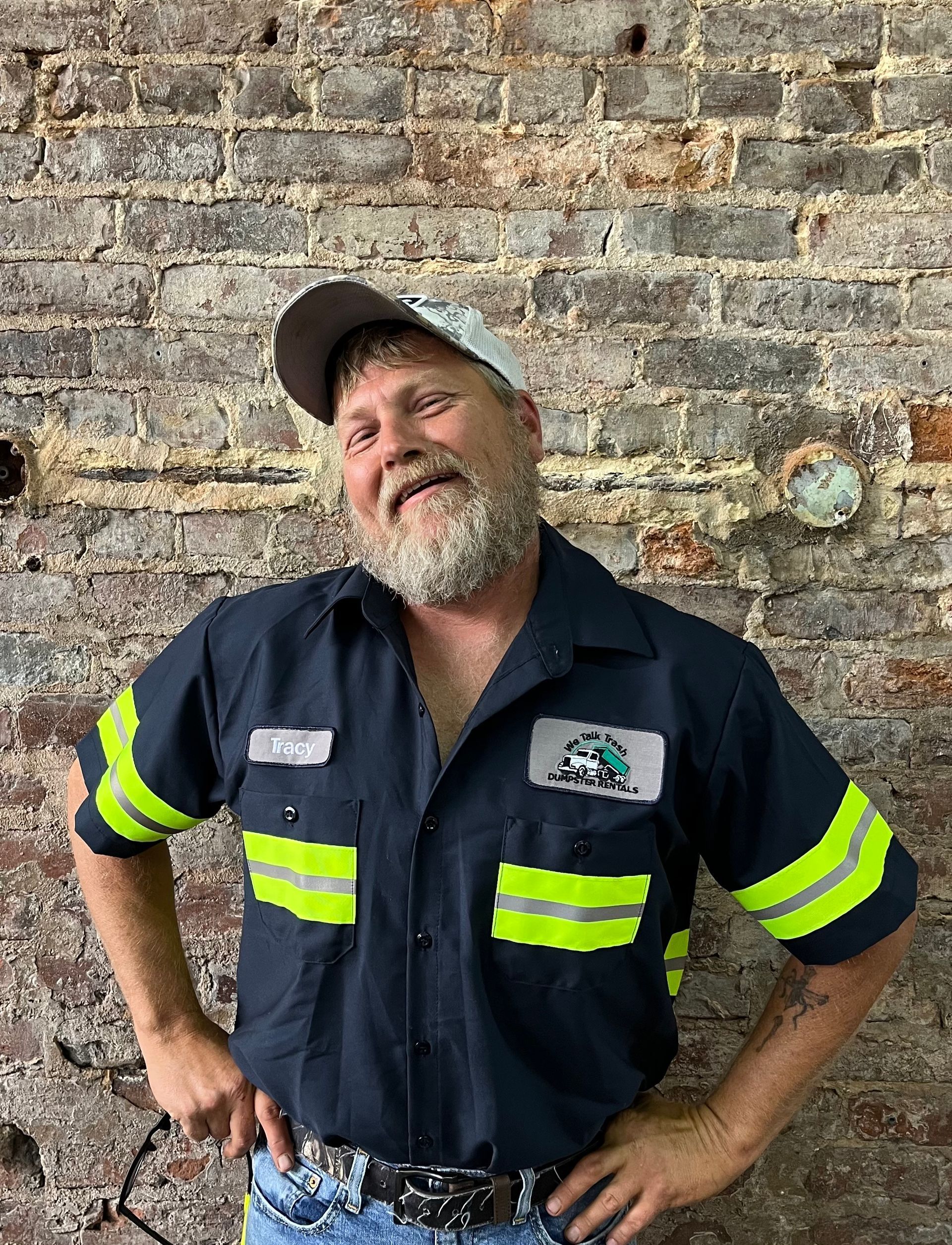 A man with a beard is standing in front of a brick wall with his hands on his hips.
