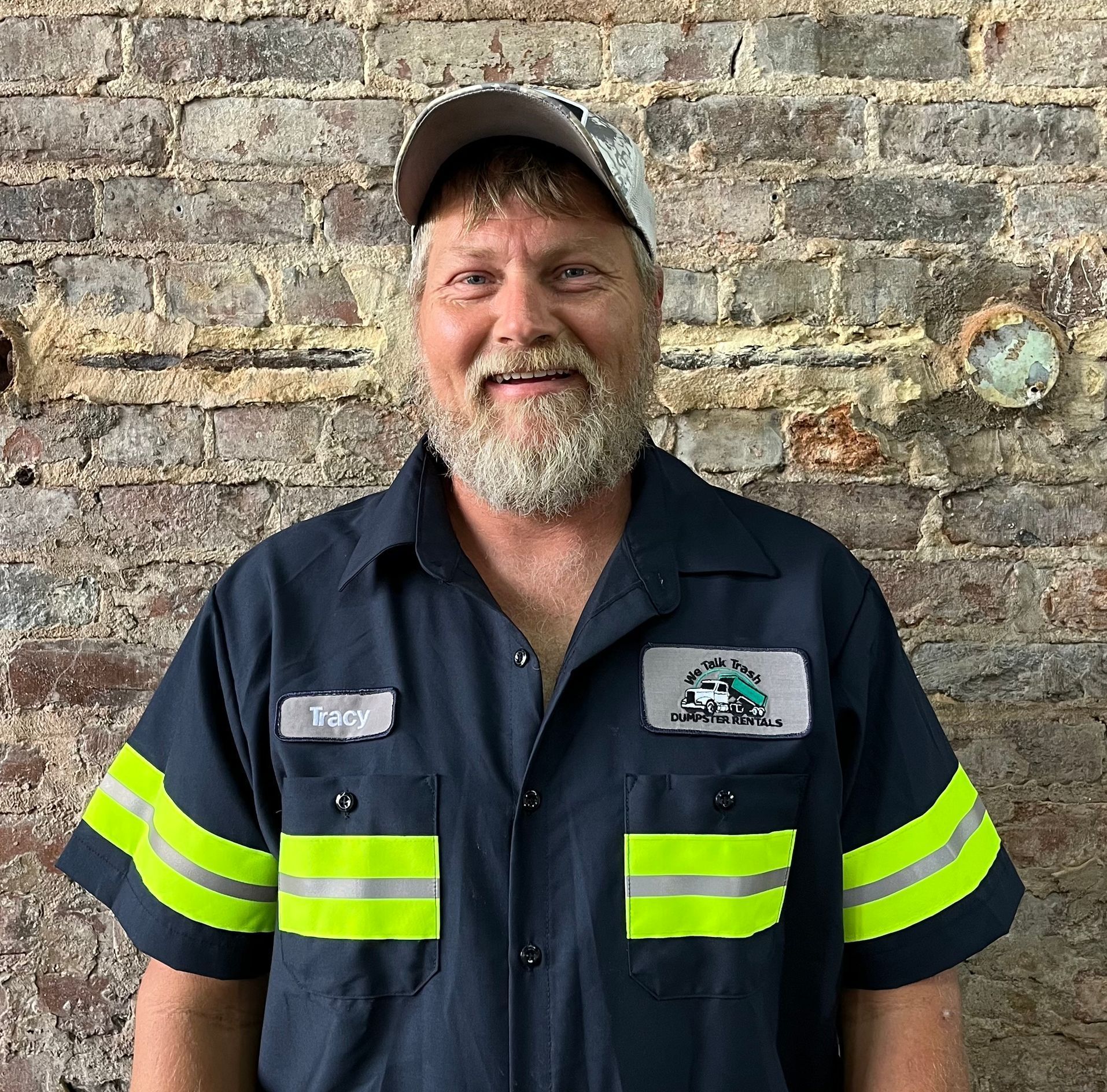 A man with a beard and a hat is standing in front of a brick wall.