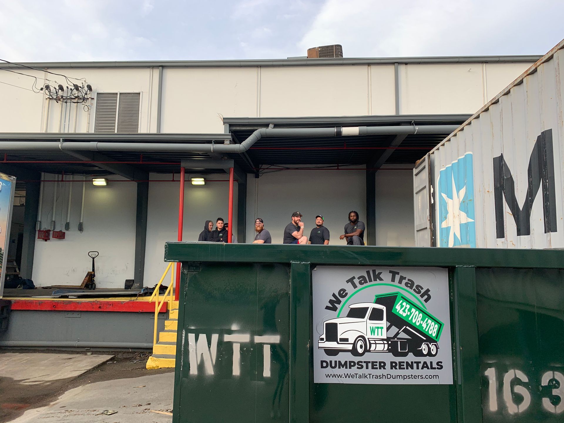 A group of people standing on top of a dumpster in front of a building.