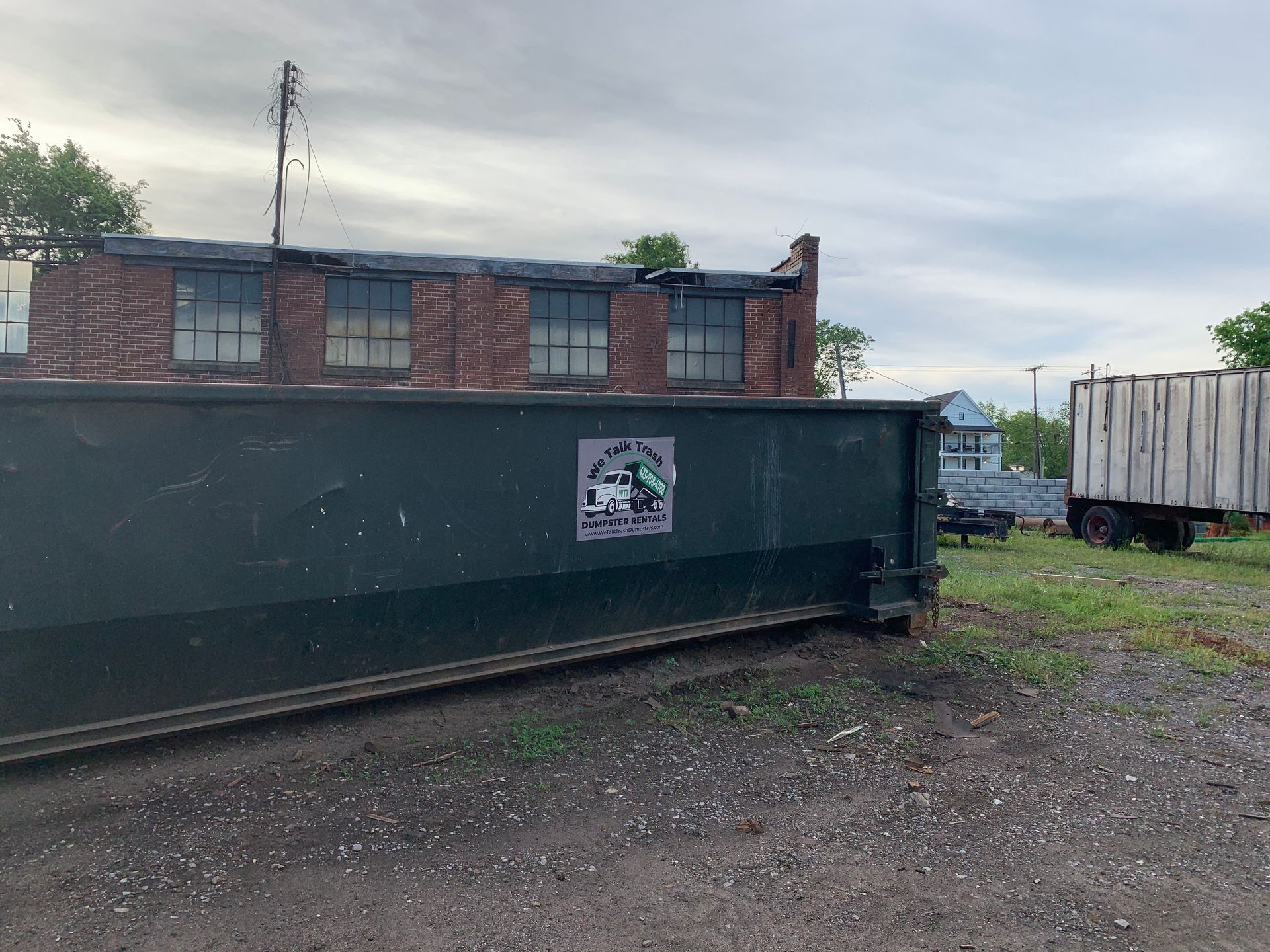 A dumpster is sitting in a gravel lot in front of a brick building.