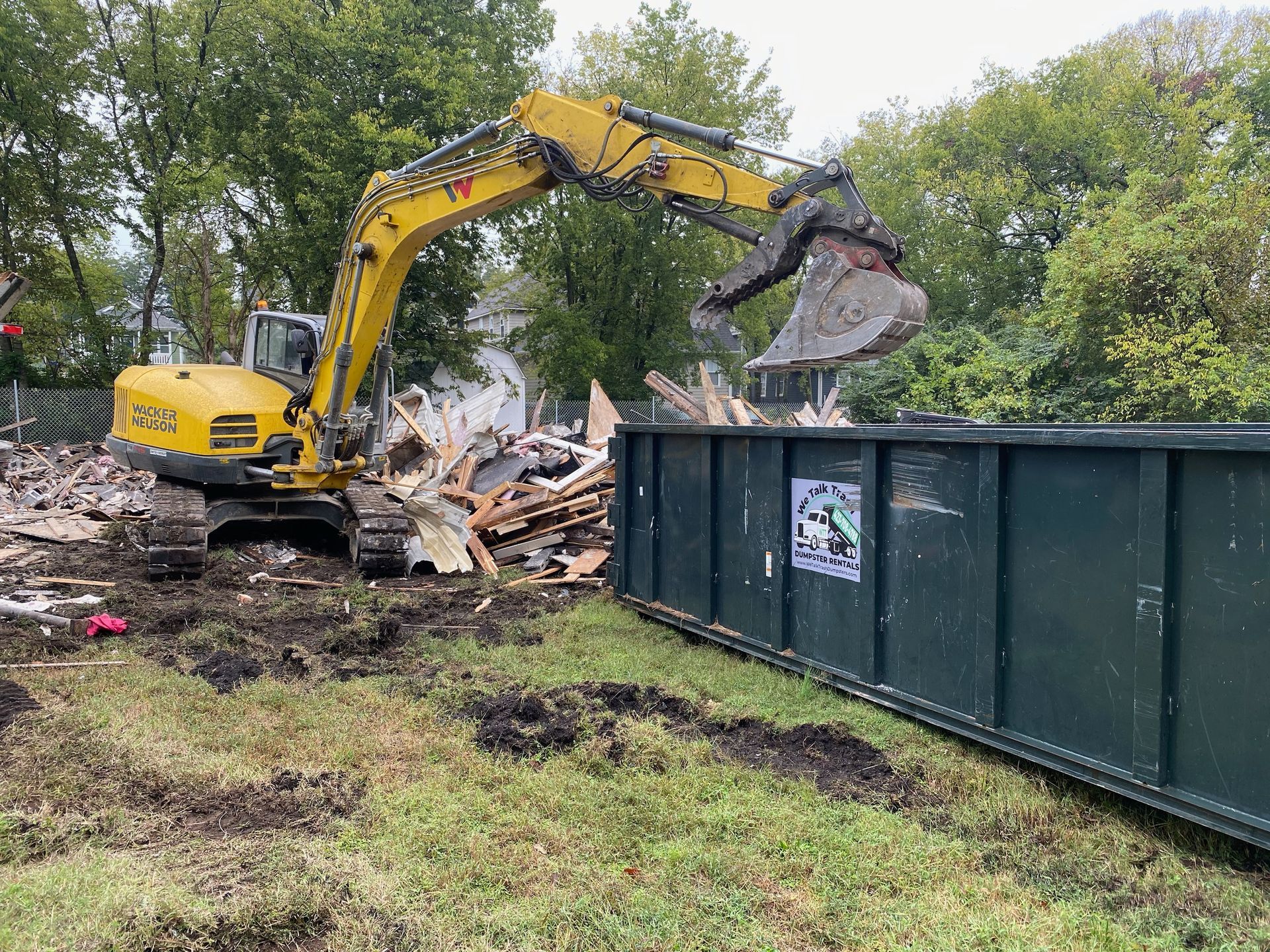 A yellow excavator is demolishing a building in a field next to a dumpster.