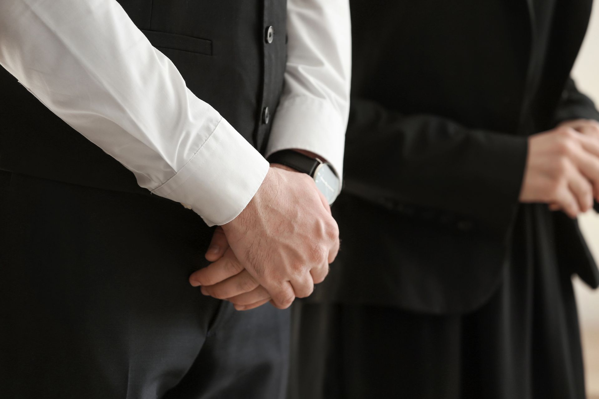 Due persone vestite con abiti formali neri e camicie bianche, in piedi con le mani giunte davanti a sé.