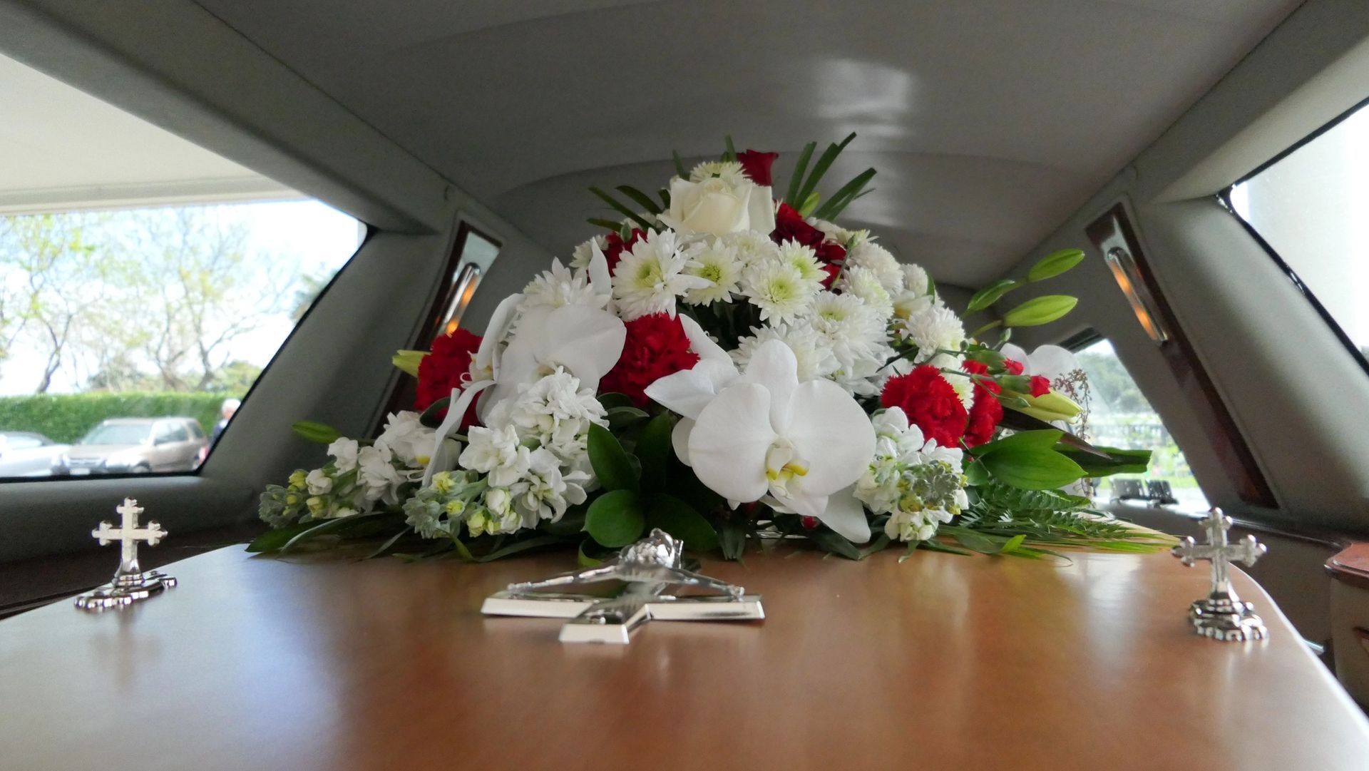 Una bara in un carro funebre adornata con un grande mazzo di fiori bianchi e rossi e tre piccole croci d'argento.