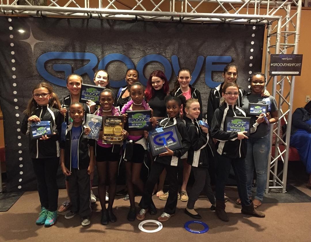 Group of smiling dancers holding trophies and medals in front of a Groove backdrop at an awards event Group of smiling dancers holding trophies and medals in front of a Groove backdrop at an awards event