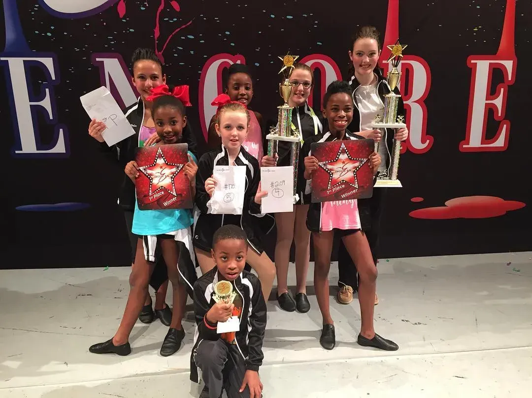 Children posing on a stage with red and black backdrop, holding awards and certificates. Children posing on a stage with red and black backdrop, holding awards and certificates.