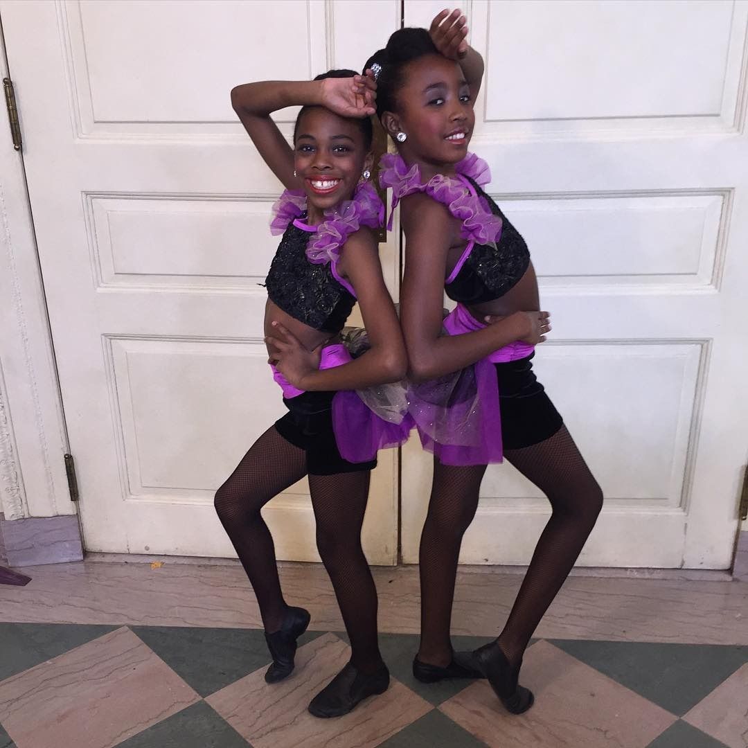 Two dancers in black and purple costumes pose back-to-back in front of white doors. Two dancers in black and purple costumes pose back-to-back in front of white doors.