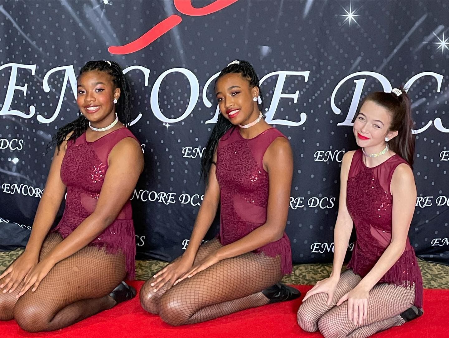Three dancers in burgundy outfits kneel on a red carpet at a formal event backdrop