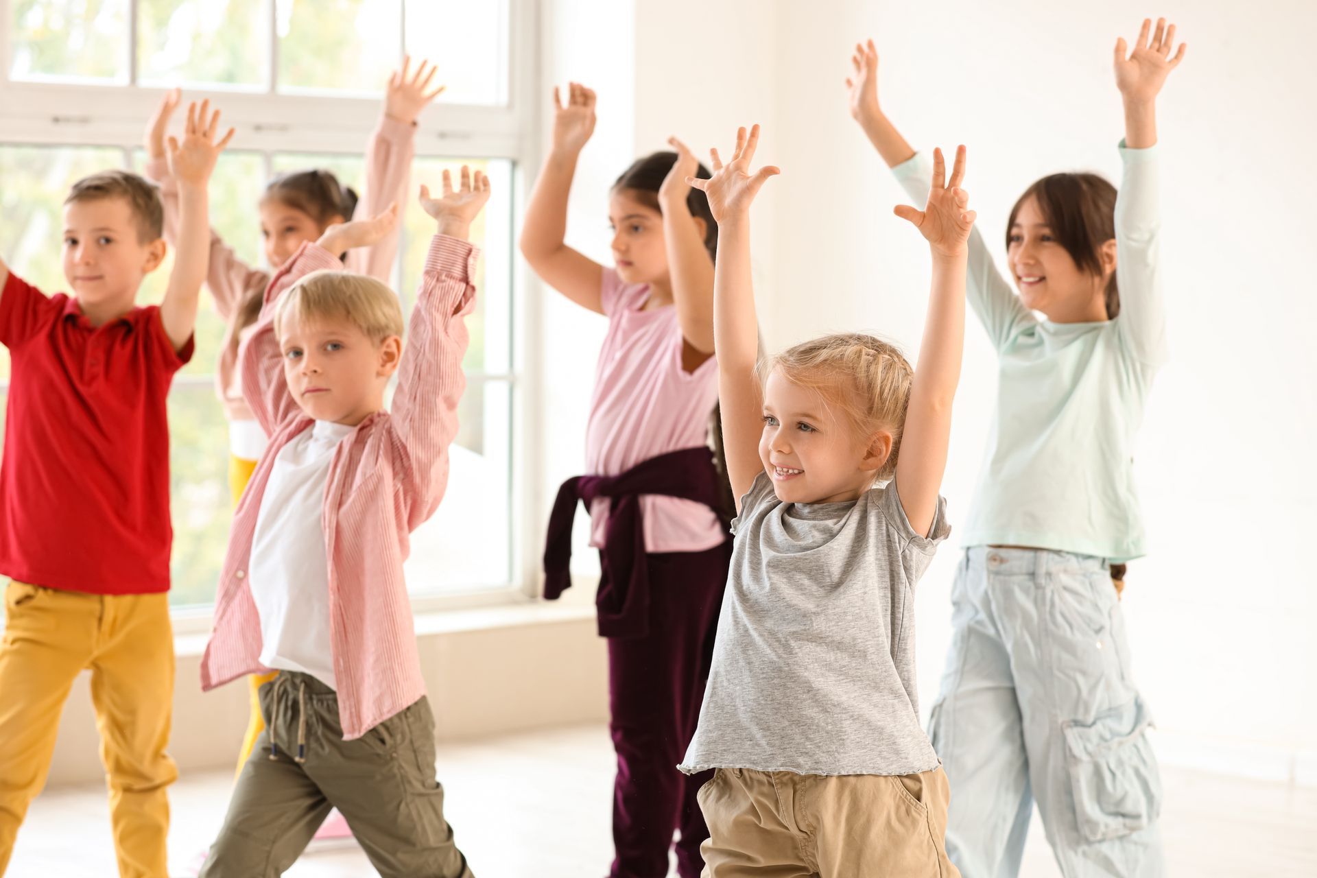 Children stretching with arms raised in a bright classroom near large windows Children stretching with arms raised in a bright classroom near large windows