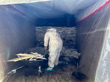 Worker in white protective suit inspecting stacked materials inside a tarped industrial container.