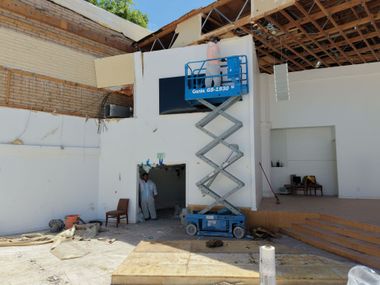Scissor lift raising workers indoors beneath a high, partially roofed ceiling under construction