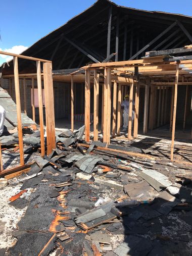 Burned wooden structure with collapsed roof and charred debris in front under bright daylight