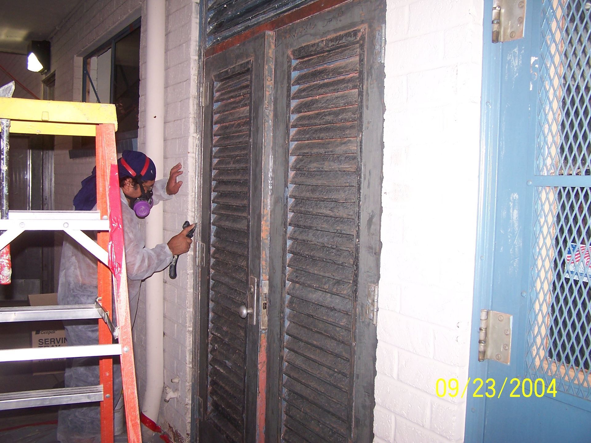 Worker painting exterior wall beside shuttered doors, with ladder and safety gear.