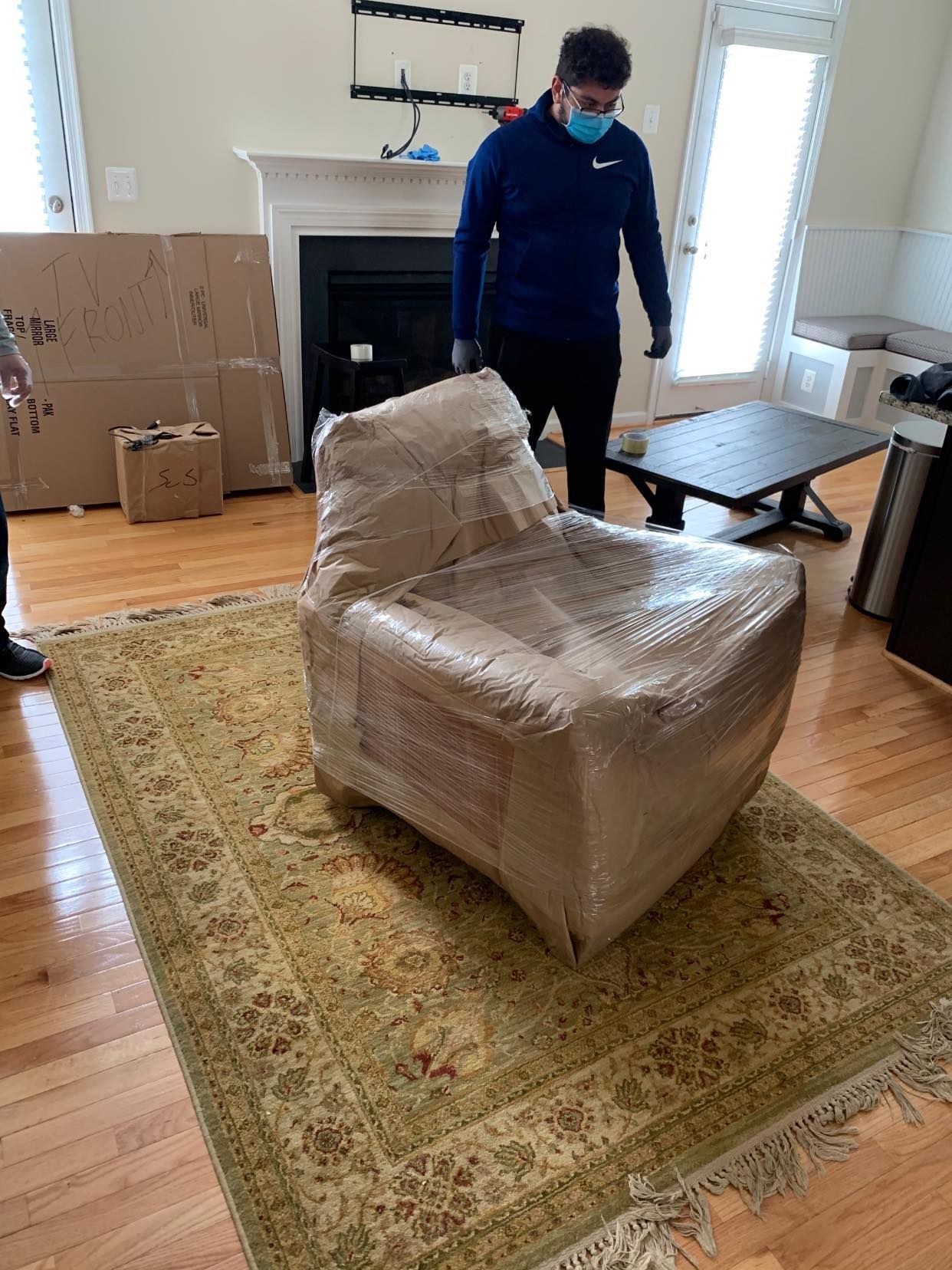 A man wearing a mask is standing next to a chair wrapped in plastic.