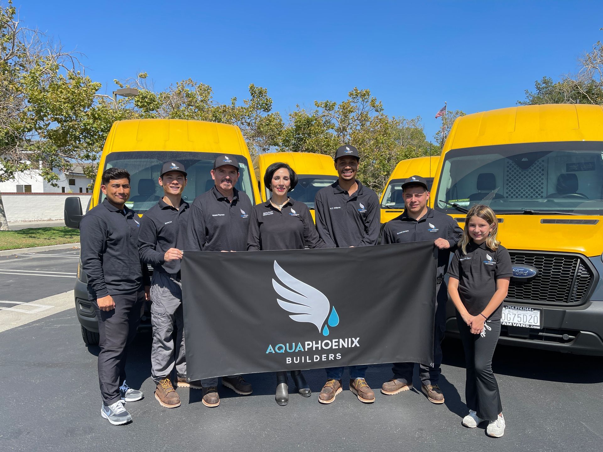 A group of people in black shirts and caps stand in front of yellow vans, holding a black flag with the Aqua Phoenix Builders logo.