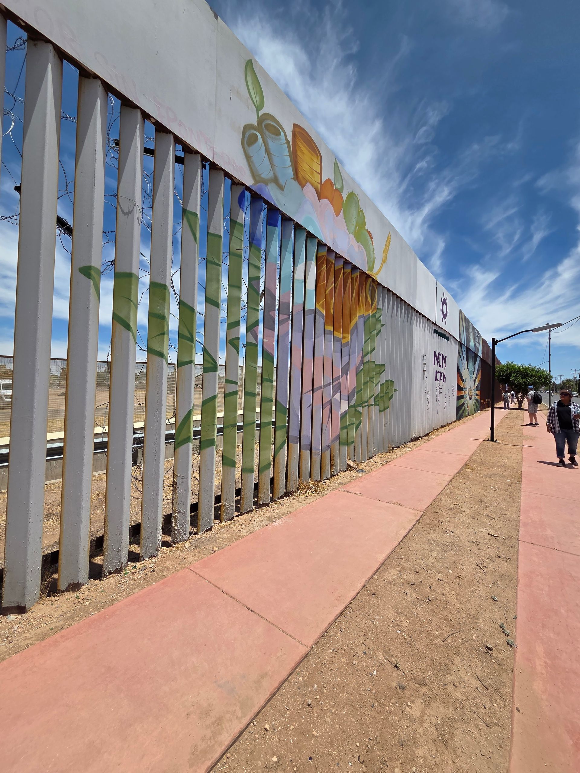 A colorful mural on a border wall with a sidewalk, blue sky, and a few people walking.