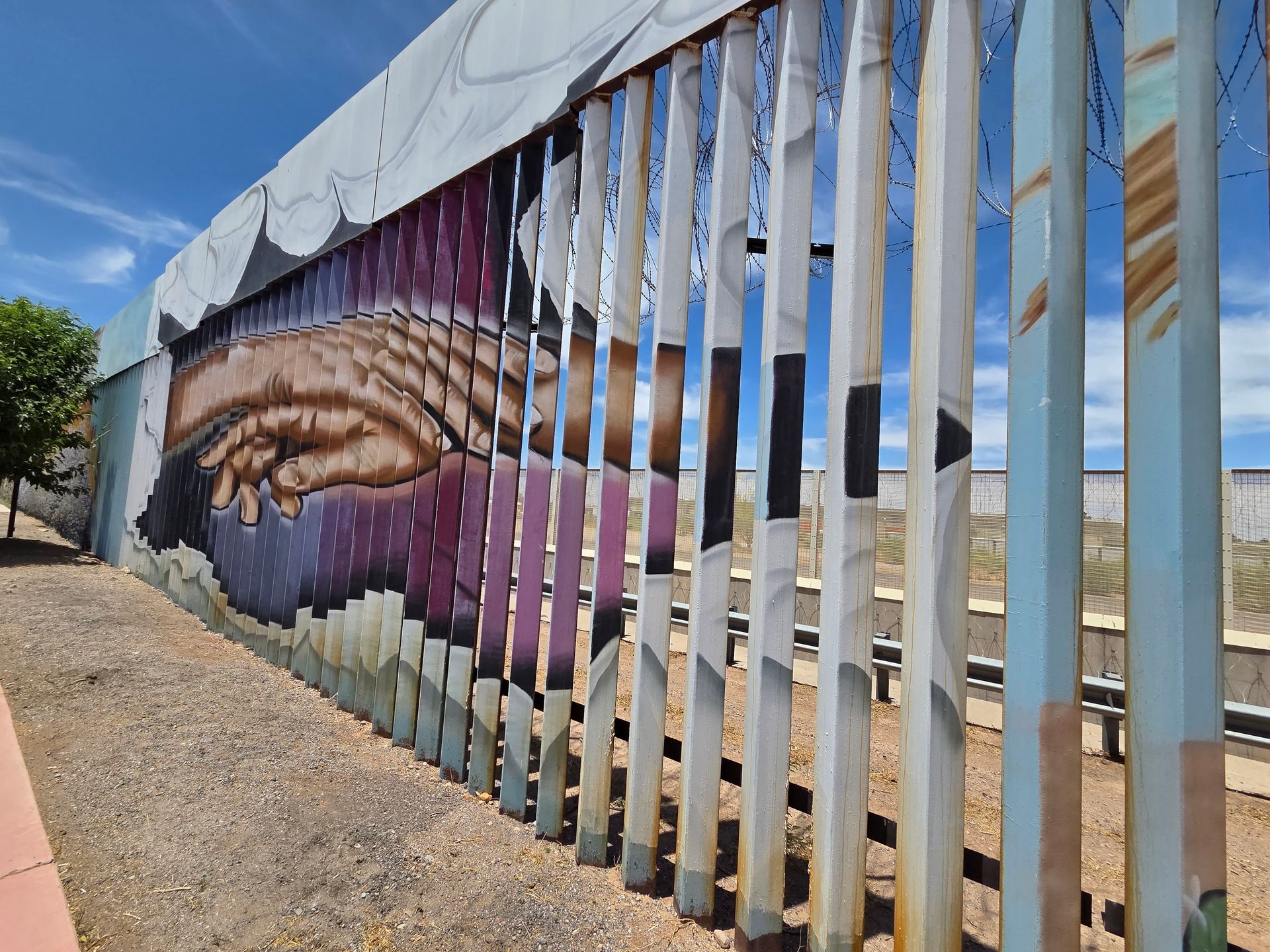 Mural of a hand reaching out, bisected by a metal border fence, with a blue sky backdrop.