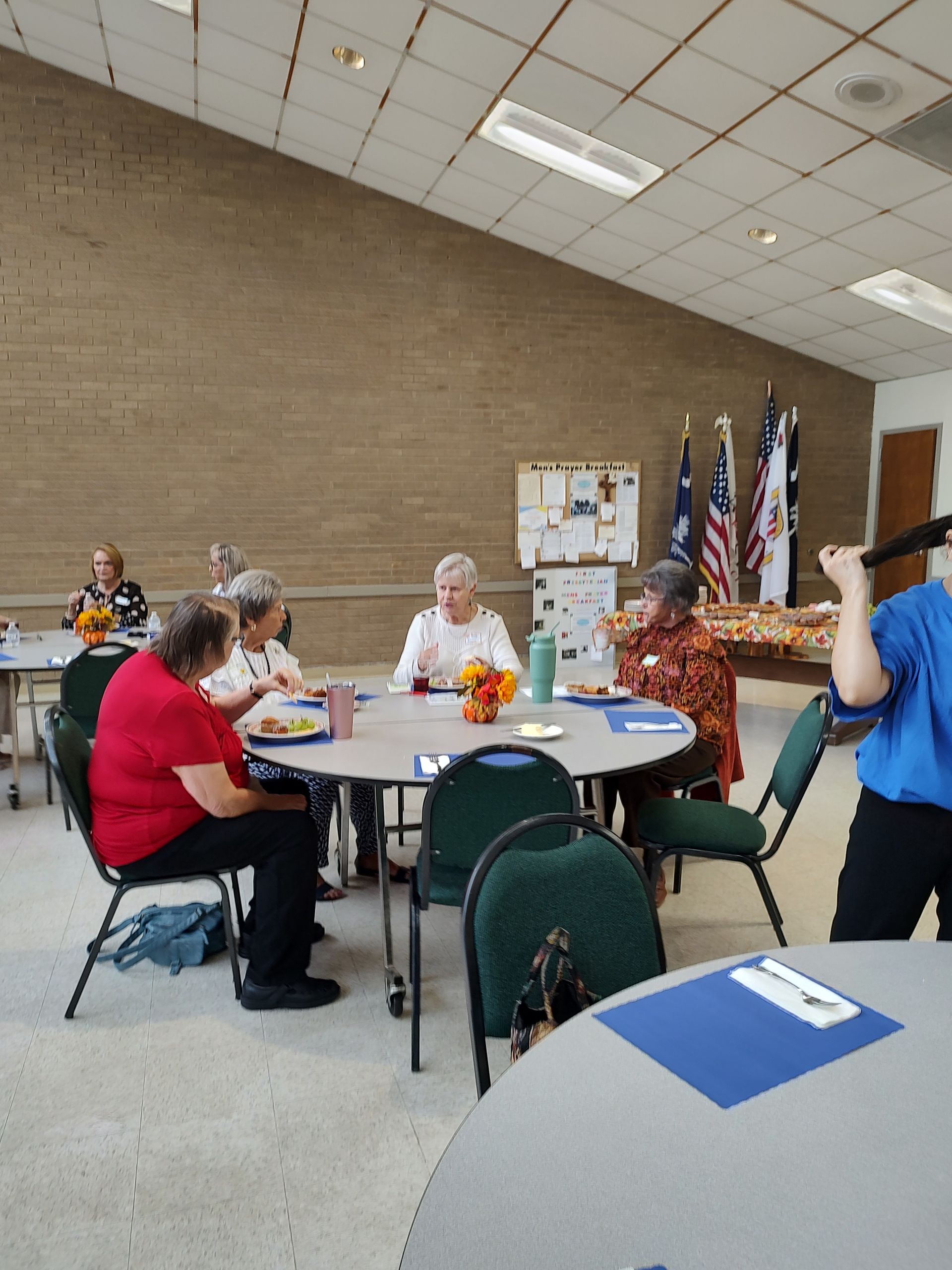 People seated at round tables, eating and socializing in a community room. Flags and a bulletin board are visible.