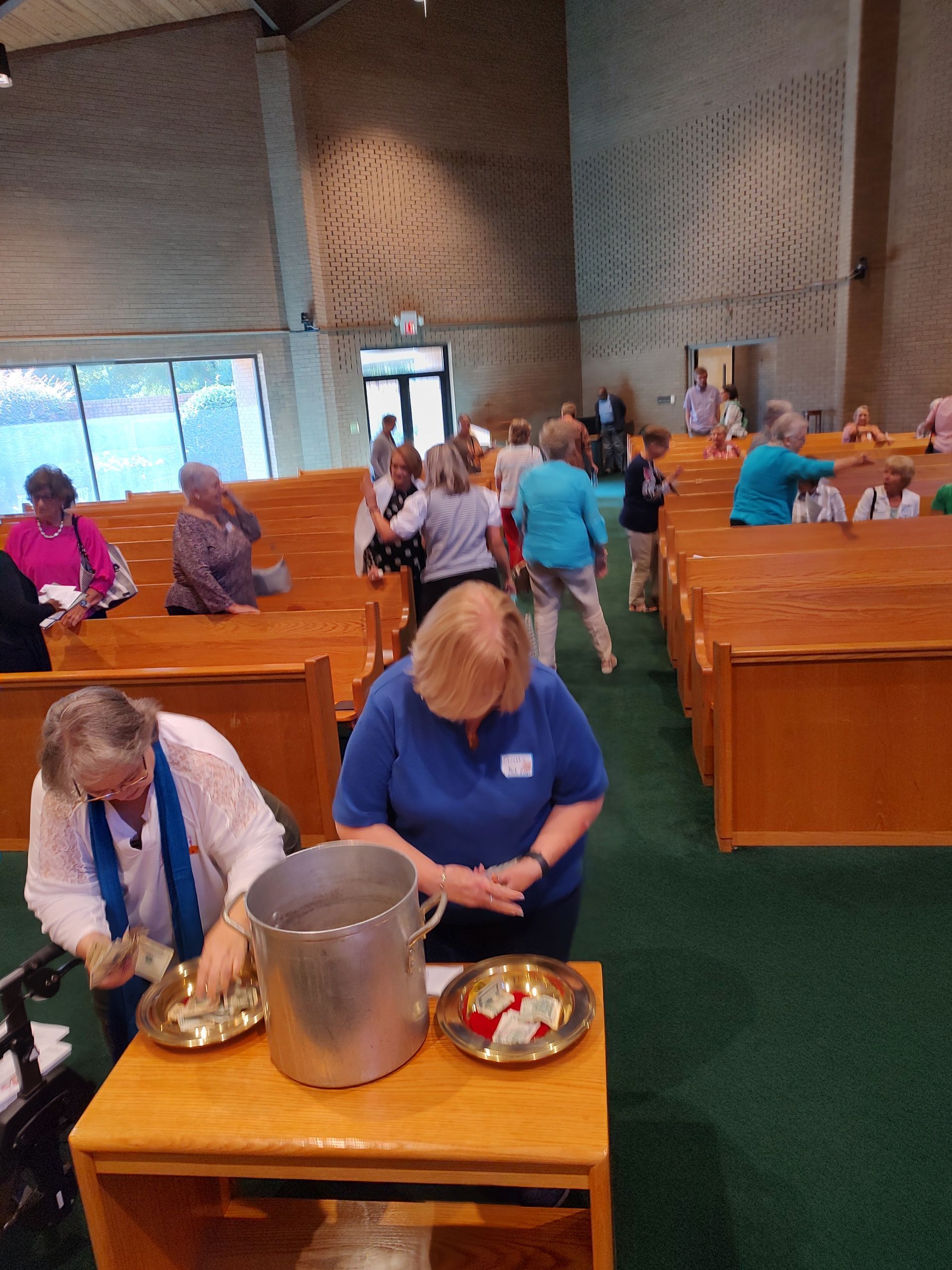 People in a church preparing or receiving communion. Two women at a table with bread and wine, others in pews.