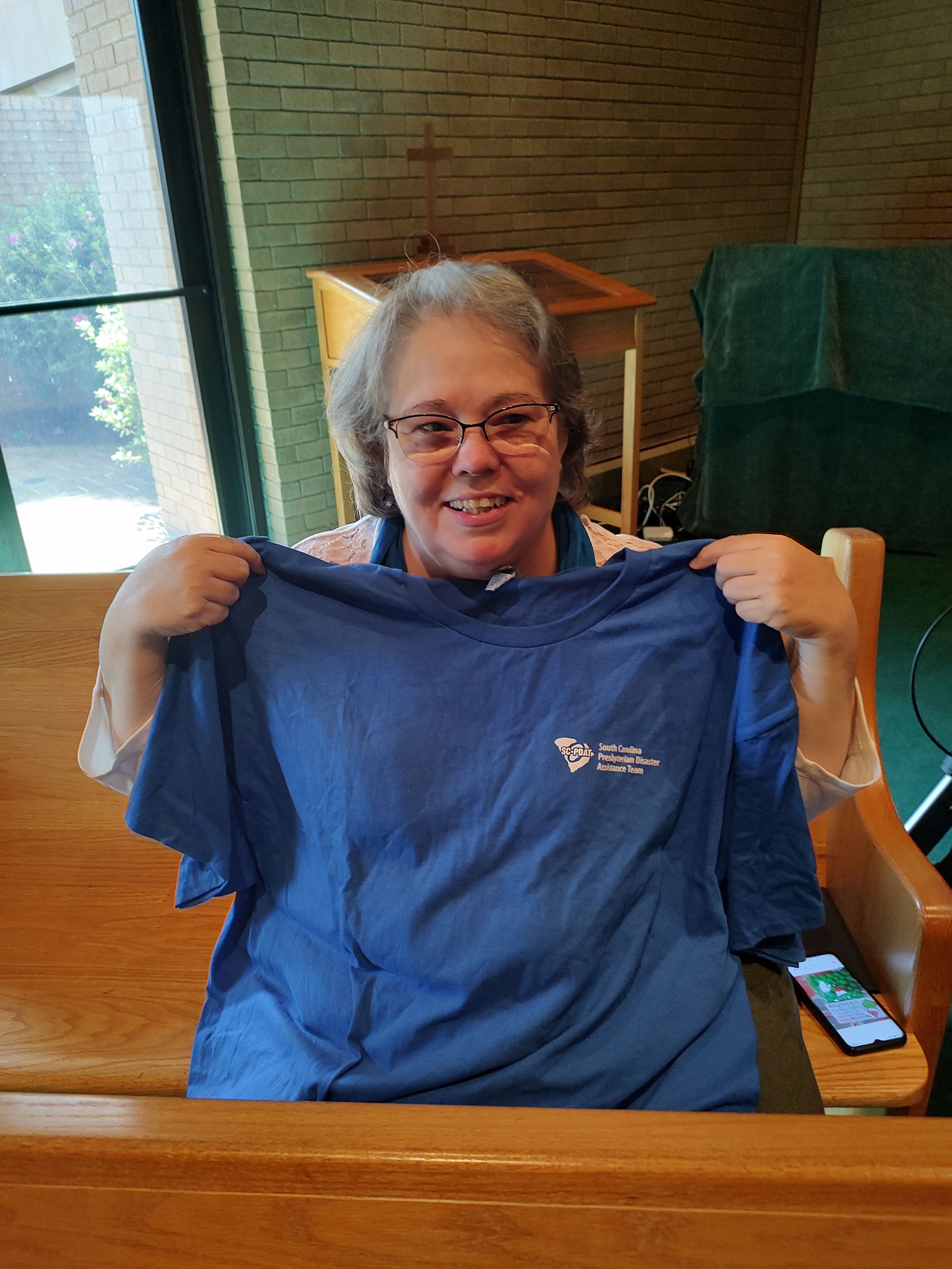 Woman in glasses holding a blue shirt with a logo, smiling in a wood-paneled room with a window.