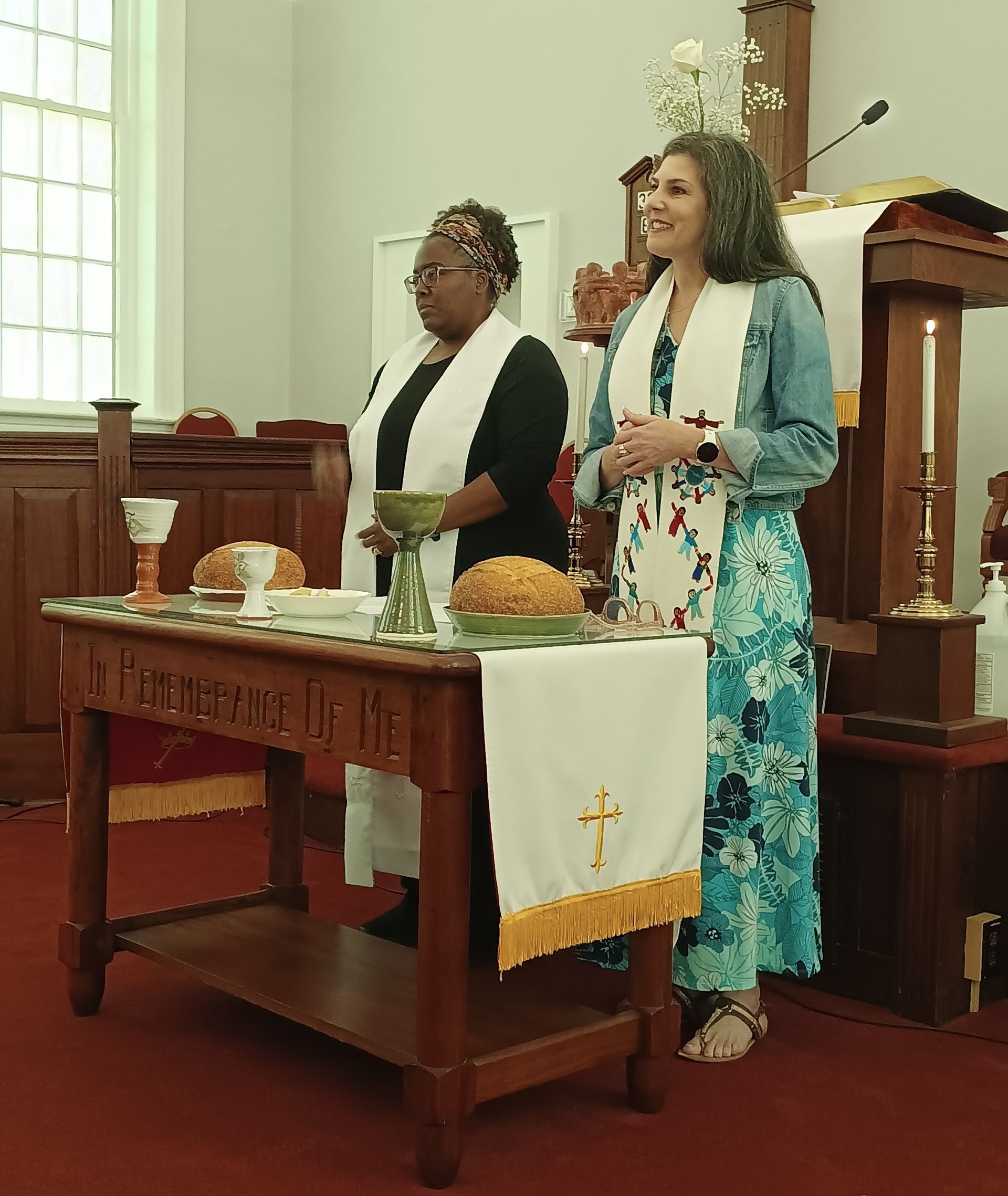 Two women standing at a church altar, preparing for communion. One wears a denim jacket, the other a black top.