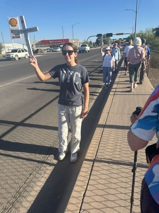 A woman holds a cross and leads a group walking on a sidewalk near a road under a sunny sky.