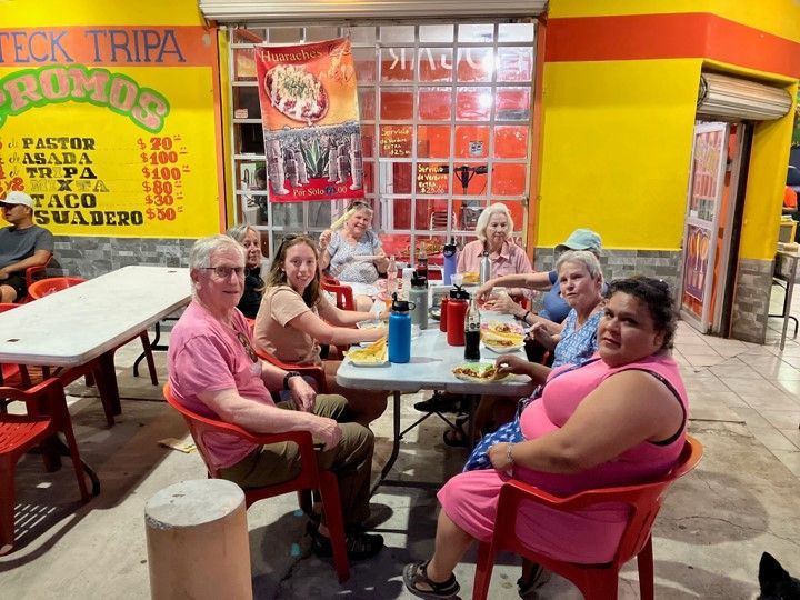 Group of people sitting at a table outside a taco restaurant; yellow walls, red chairs, food visible.