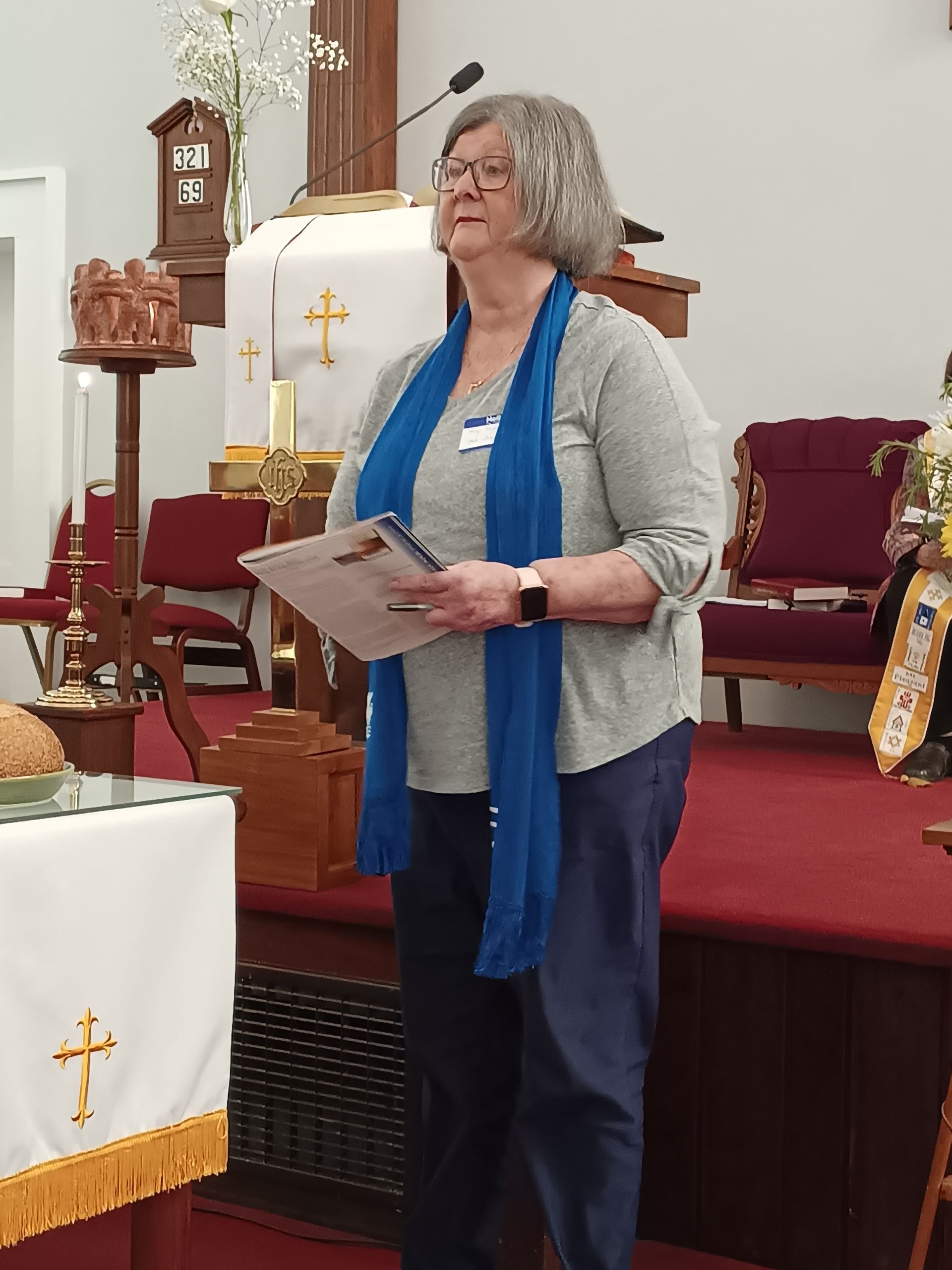 Woman in a church holding papers, wearing a blue scarf. She stands in front of a cross.